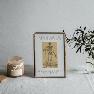 May include: A framed print with a gold frame and a black and white illustration of a skeleton. The text on the print reads "We'll lay here for years or for hours so long, we'd become the flowers."