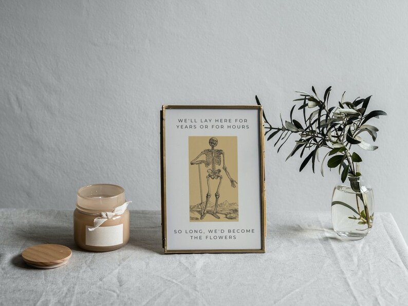May include: A framed print with a gold frame and a black and white illustration of a skeleton. The text on the print reads "We'll lay here for years or for hours so long, we'd become the flowers."