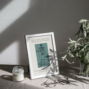 May include: A white framed print with a black and white illustration of a skeleton standing with a cane. The text on the print reads "We'll lay here for years or for hours. So long, we'd become the flowers."