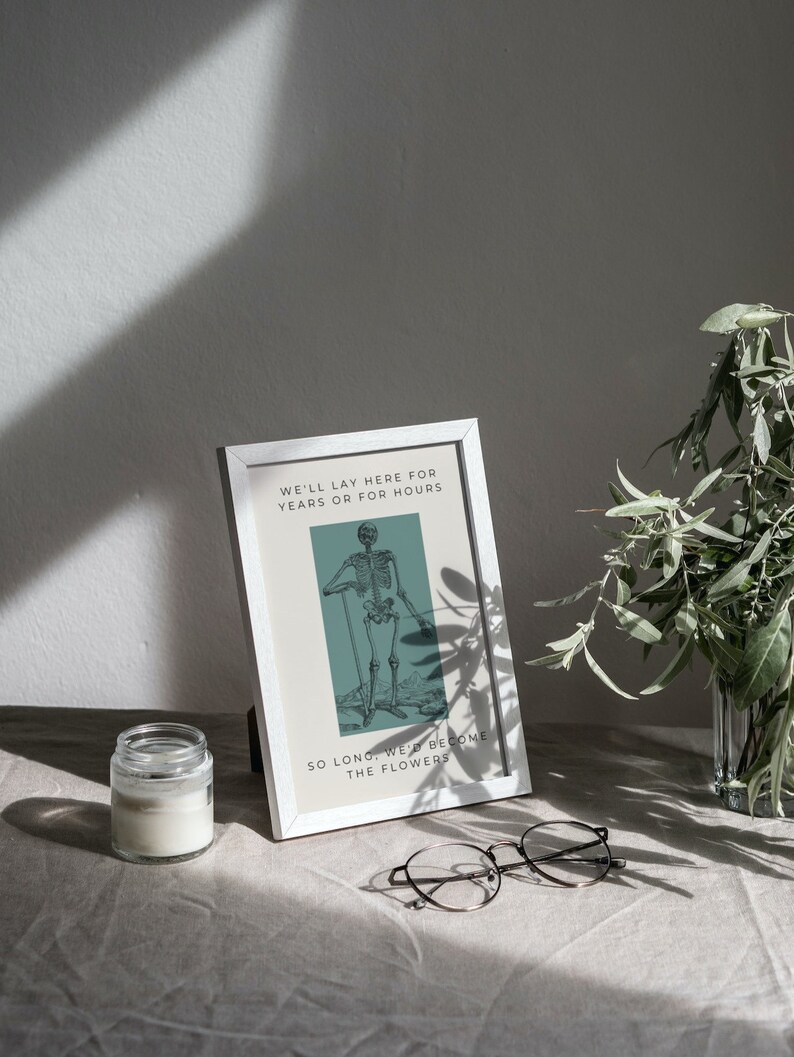 May include: A white framed print with a black and white illustration of a skeleton standing with a cane. The text on the print reads "We'll lay here for years or for hours. So long, we'd become the flowers."