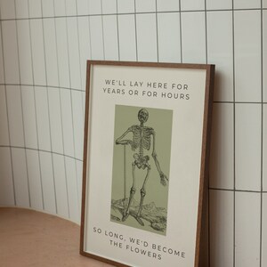 May include: A framed print with a green background featuring a black and white skeleton illustration. The text reads "We'll lay here for years or for hours. So long, we'd become the flowers."