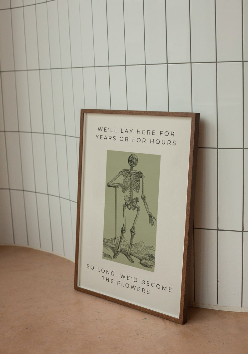 May include: A framed print with a green background featuring a black and white skeleton illustration. The text reads "We'll lay here for years or for hours. So long, we'd become the flowers."