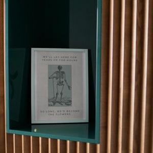 May include: A framed print with a black and white illustration of a skeleton standing with a stick. The text on the print reads "We'll lay here for years or for hours. So long, we'd become the flowers."