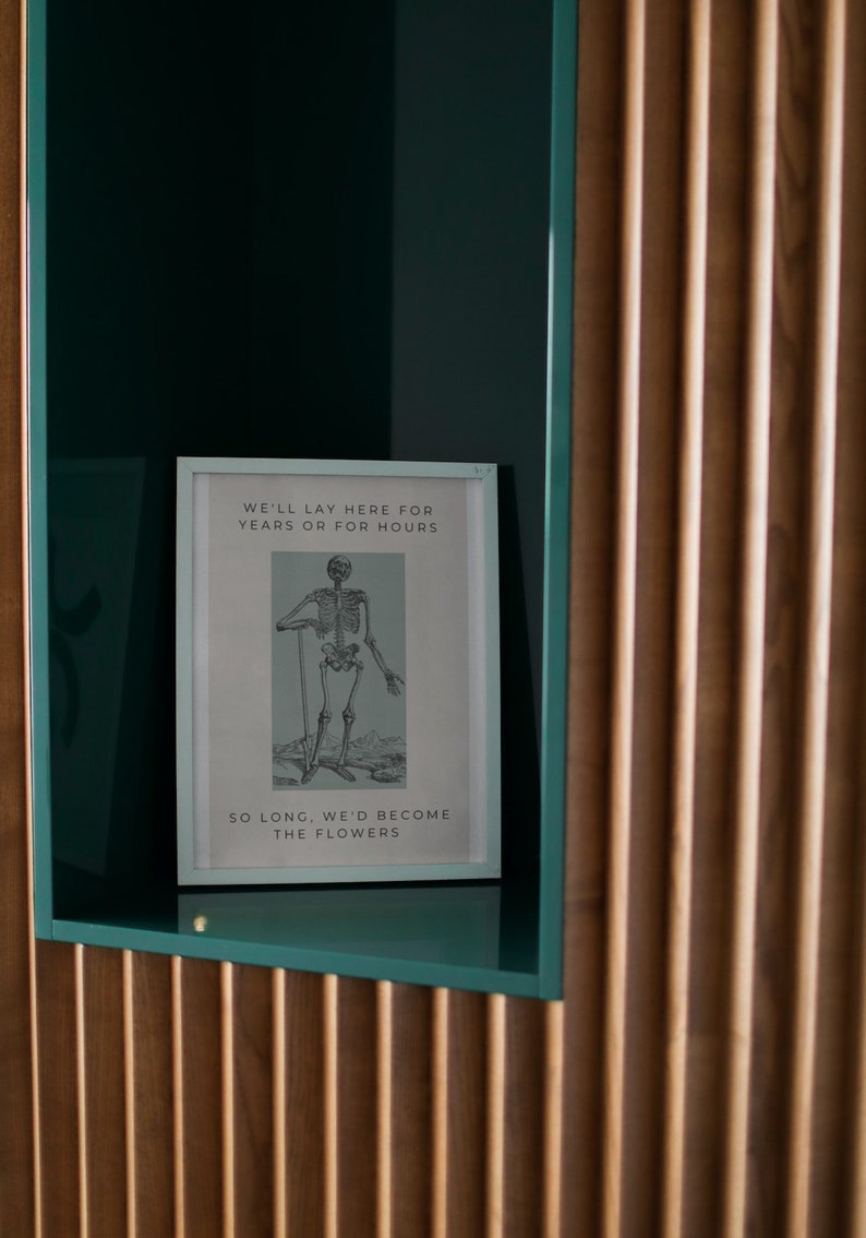May include: A framed print with a black and white illustration of a skeleton standing with a stick. The text on the print reads "We'll lay here for years or for hours. So long, we'd become the flowers."