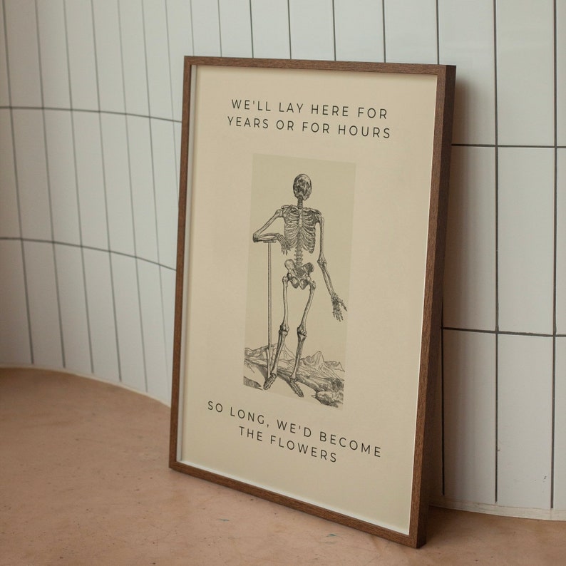 May include: A framed print featuring a black and white illustration of a skeleton standing with a stick in its hand. The text on the print reads "We'll lay here for years or for hours, so long we'd become the flowers."