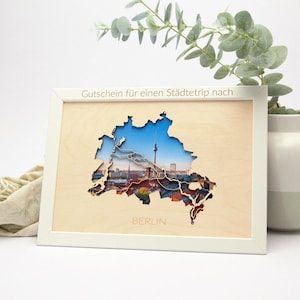 Op de afbeelding: Een wit ingelijst houten uitsnijding van de stad Berlijn, Duitsland. De uitsnijding is een kaart van de stad met een foto van de skyline van de stad op de achtergrond. De tekst "Gutschein für einen Städtetrip nach BERLIN" is op de lijst gedrukt.