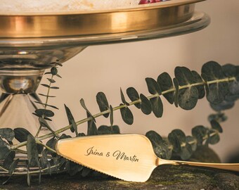 May include: A gold cake server with the inscription "Irina & Martin" on a mossy rock with greenery in the background.