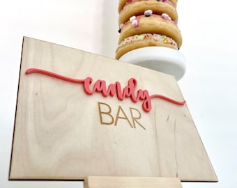 May include: A wooden sign with the words "candy bar" in pink lettering on a wooden easel. The sign is in front of a tower of donuts with pink and white frosting and sprinkles.