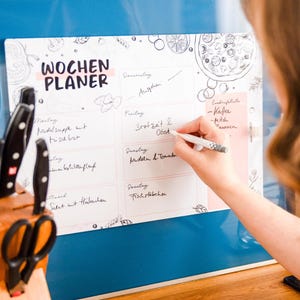 May include: A white weekly planner with the words "WOCHEN PLANER" in black. The planner has a pink border and is being written on with a pen. A wooden knife block with black knives and scissors is in the foreground.