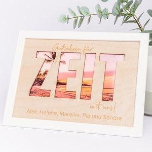 Puede incluir: Un letrero de madera enmarcado en blanco con un recorte de la palabra "ZEIT" (alemán para "tiempo") con una escena de playa visible a través de las letras. El letrero dice "Gutschein für ZEIT mit uns!" (alemán para "Vale por tiempo con nosotros!") y "Alex, Helene, Mareike, Pia und Sandra".