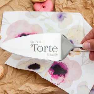 Könnte beinhalten: Ein silberner Tortenheber mit einem gezackten Rand und dem Text "KAUM IN TORTE ZU FASSEN" auf der Klinge. Der Heber liegt auf einer weißen Serviette mit einem Blumenmuster.