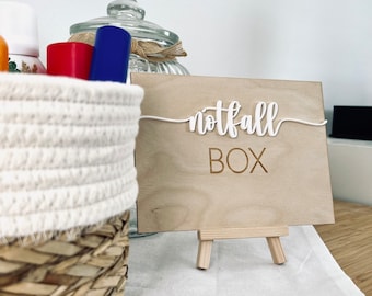 May include: A wooden sign with the text "notfall BOX" on a small easel. The sign is sitting on a white surface next to a wicker basket filled with various items.