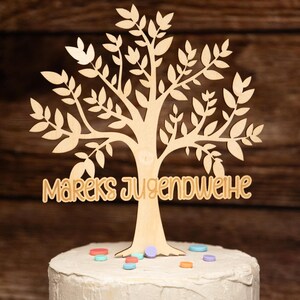 May include: A wooden tree cake topper with the words "Mareks Jugendweihe" on the trunk. The tree is brown and decorated with colourful sprinkles.