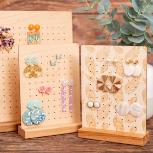 Puede incluir: Tres soportes de exhibición de aretes de madera con varios estilos de aretes. Los soportes están hechos de madera natural y tienen un aspecto rústico. Los aretes están hechos de diferentes materiales, incluyendo metal, plástico y madera.