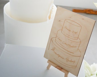 May include: A wooden sign with a hand-drawn illustration of a three-tiered wedding cake. The sign reads "Salted - Maple, Lemon - Coconut & Strawberry, Mascarpone - Bailey's".