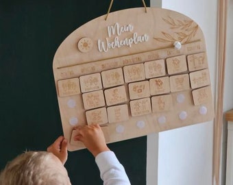 May include: A wooden weekly planner with the text "Mein Wochenplan" in white lettering. The planner has a light wood finish and features a sun, a flower, and leaves. The planner is hung on a wall and a child is interacting with it.