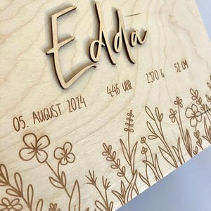 May include: A wooden plaque with the name "Edda" engraved in cursive script. The plaque also includes the date "05. August 2014", the time "4:46 Uhr", the weight "2370 g", and the length "51 cm". The plaque is decorated with a floral design.