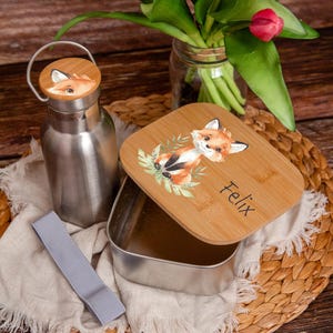 Könnte beinhalten: Eine Edelstahl-Wasserflasche und eine Metall-Lunchbox mit Bambusdeckel. Der Deckel zeigt eine Aquarell-Illustration eines Fuchses und den Namen "Felix". Ein graues Gummiband und ein geflochtener Untersetzer sind ebenfalls sichtbar.