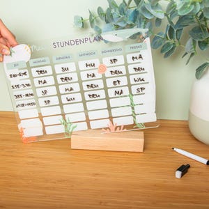 May include: A clear acrylic weekly planner with the German word "STUNDENPLAN" at the top. The planner is decorated with sea-themed illustrations and sits on a wooden block. A hand holds a decorative sticker.