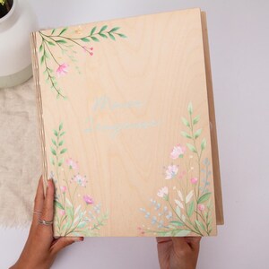 Op de afbeelding: Een lichtgekleurde houten kist met bloemwerk. De kist heeft handgeschilderde roze en blauwe bloemen met groene bladeren. De tekst "Mama, Longing" is in lichtblauw schrift over de voorkant van de kist geschreven.