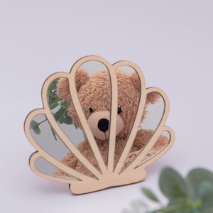 Op de afbeelding: Een houten spiegel in de vorm van een schelp met een teddybeer die erin wordt weerspiegeld. De spiegel is gemaakt van licht hout en heeft een natuurlijke afwerking.
