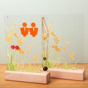Peut inclure: Présentoir à bijoux en acrylique transparent avec un motif floral peint. Le présentoir comprend des boucles d'oreilles orange et un collier doré. La base est en bois clair. Le design comprend des éléments floraux orange, jaunes, verts et rouges.