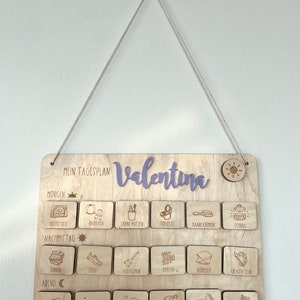 May include: A wooden daily schedule board with the name "Valentina" written in purple. The board is divided into three sections: morning, afternoon, and evening. Each section has a row of icons representing different activities, such as getting dressed, brushing teeth, playing, and reading.