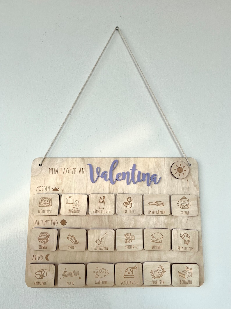 May include: A wooden daily schedule board with the name "Valentina" written in purple. The board is divided into three sections: morning, afternoon, and evening. Each section has a row of icons representing different activities, such as getting dressed, brushing teeth, playing, and reading.