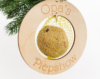 Pode incluir: Um ornamento de comedouro para pássaros de madeira com o texto "Opa's Piepshow" e uma bola de sementes para pássaros em rede pendurada dentro.