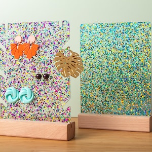 May include: Two glittery acrylic earring displays with wooden bases. One display holds orange, brown, and blue earrings. The other is a solid glitter panel. The displays are on a light green background.