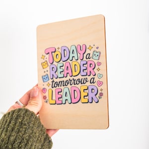 Könnte beinhalten: Rechteckige Holzplakette mit dem Text "Today a Reader, Tomorrow a Leader" in bunten Buchstaben. Das Design umfasst Sterne, Herzen und Buchillustrationen. Die Plakette wird von einer Person gehalten.
