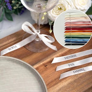May include: A wine glass with a white ribbon tied around the stem, with the name "SOFIA" printed on the ribbon. Additional ribbons with names are on a wooden surface. A colour chart of ribbons is in the background.