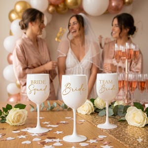 Może przedstawiać: Trzy białe kieliszki do wina ze złotym napisem na drewnianym stole. Lewy kieliszek ma napis Bride Squad, środkowy Bride, a prawy Team Bride. Kieliszki są otoczone białymi różami i konfetti.
