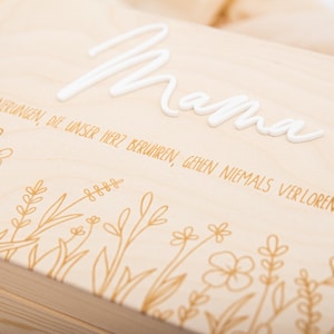 May include: A wooden box with a white inscription that reads "Mama". The box is decorated with a gold floral design and the text "Erinnerungen, die unser Herz berühren, gehen niemals verloren".
