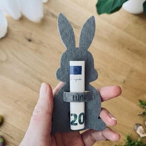 Peut inclure: Un porte-monnaie en forme de lapin en feutre gris avec un billet enroulé. Le billet a un motif bleu et jaune et le chiffre 20 en vert. Le support porte le nom "LUNA".