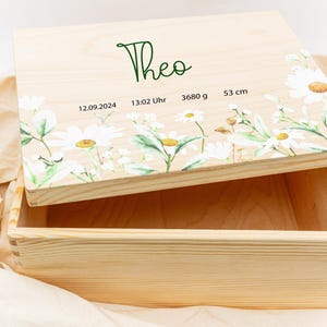 Può includere: Scatola portaoggetti in legno con finitura in legno chiaro e un design floreale sul coperchio. Il coperchio presenta un motivo di margherite bianche con foglie verdi e il nome "Theo" scritto in corsivo verde. La scatola include anche la data "12.09.2024", l'ora "13:02 Uhr", il peso "3680 g" e la lunghezza "53 cm".