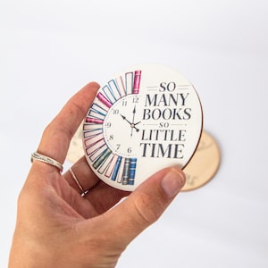 Könnte beinhalten: Ein runder Untersetzer mit einem Zifferblatt-Design, das farbenfrohe Buchrücken am Rand zeigt. Der Text lautet "SO MANY BOOKS SO LITTLE TIME". Die Zeiger der Uhr zeigen auf 10:10. Der Untersetzer wird in einer Hand gehalten.