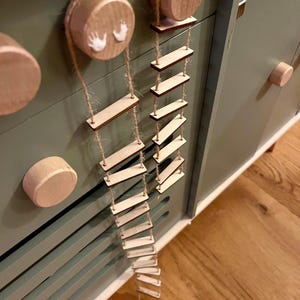Könnte beinhalten: Holzspielzeugleiter mit kleinen rechteckigen Stufen, die an Schnüren aufgehängt sind. Die Leiter ist an einem Holzzylinder mit weißen Handabdrücken befestigt. Die Leiter hängt an einem grünen Schrank mit Holzknöpfen.