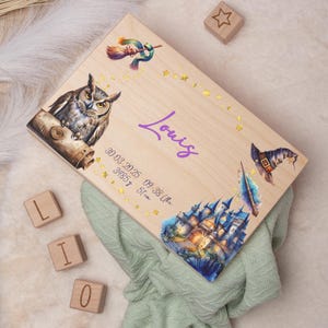 Puede incluir: Una caja de madera para recuerdos con un tema mágico de Harry Potter. La caja presenta un búho morado, un castillo, una varita mágica y un sombrero de bruja. La caja está personalizada con el nombre "Louis" y la fecha "30.03.2025 09:38 Uhr" junto con el peso y la longitud del bebé.