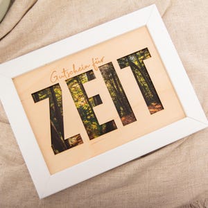May include: A white framed wooden sign with the word "ZEIT" cut out, revealing a forest scene. The text "Gutschein für" is above the cut-out letters. The sign is on a neutral-colored fabric.