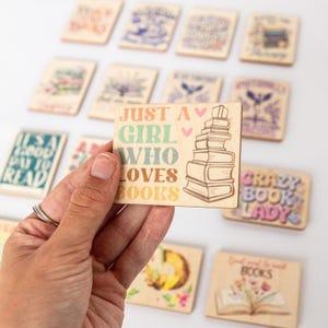 May include: Wooden rectangular magnet with the text "JUST A GIRL WHO LOVES BOOKS" in pastel colors, with a stack of books illustration. Other magnets with book-related quotes are in the background.