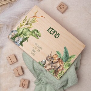 May include: Wooden keepsake box with a dinosaur theme. The box is decorated with a green Tyrannosaurus Rex, a brown Triceratops, and green leaves. The box is personalized with the name "Keno" and the date "01.03.2025" along with the time "02:45 Uhr", weight "3250 g", and length "50 cm".