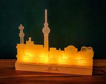 May include: A lit-up Berlin skyline silhouette lamp. The lamp is made of a light-colored material and sits on a wooden base. The word "Berlin" is written on the base. The lamp emits a warm, yellow glow, highlighting the city's iconic landmarks.