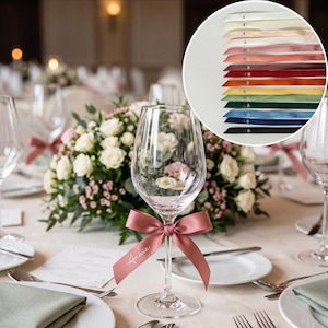 Può includere: Un calice da vino in vetro trasparente con un nastro di raso rosa polvere annodato a fiocco sullo stelo. Il nastro è personalizzato con il nome Annie in corsivo bianco. Un riquadro mostra una palette di quindici diversi nastri di raso.