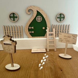 以下が含まれることがあります： 緑色のドア、2つの丸い窓、はしご、フェンス、郵便受け、「Tipsi」と書かれた看板を備えた、気まぐれな木製の妖精のドアセット。 セットには、ウェルカムマットと装飾的な足跡が含まれています。