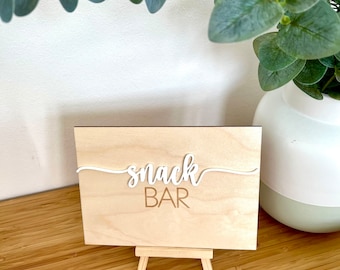 May include: A wooden sign with the words "snack bar" written in white cursive letters on a wooden easel. The sign is on a wooden surface.
