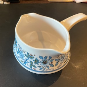 May include: White ceramic gravy boat with a blue and green floral design. The boat has a handle and a wide, rounded base.