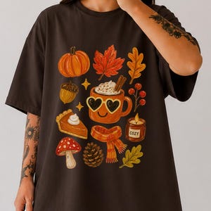 Könnte beinhalten: Ein dunkelbraunes Kurzarm-T-Shirt mit einem Grafikdruck von herbstlichen Motiven, darunter ein Kürbis, ein Ahornblatt, eine Eichel, ein Stück Kuchen, ein Pilz, ein Tannenzapfen, ein Schal, eine Kerze und eine lächelnde Tasse mit Herzbrille.