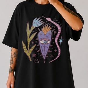 Könnte beinhalten: Ein schwarzes Kurzarm-T-Shirt mit einer zentralen Grafik eines violetten Herzens mit einem Auge, umgeben von einer blauen Blume, einer rosa Schlange und kleinen Sternen. Der Text "wild at heart" ist in kleinen weißen Buchstaben unter dem Herz gedruckt.