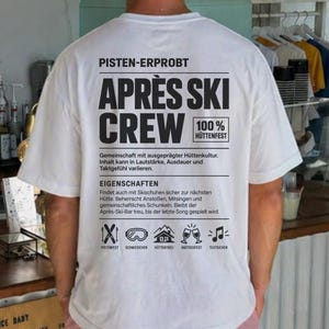 May include: A white t-shirt with black text on the back that reads APRÈS SKI CREW, PISTEN-ERPROBT, 100% HÜTTENFEST, and EIGENSCHAFTEN, along with small icons of skis, goggles, a mountain, glasses, and a musical note, paired with pink shorts.